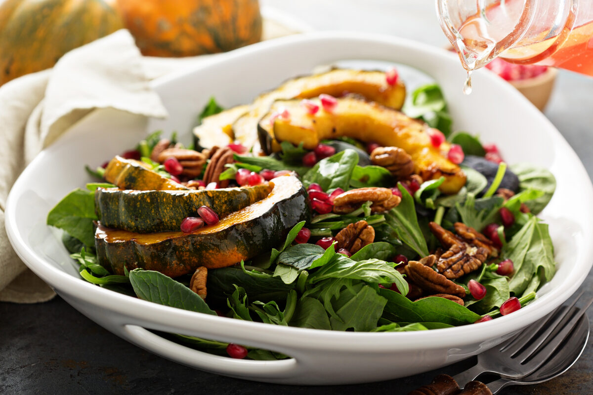 A white bowl filled with roasted acorn squash, spinach, and pomegranates with Acorn squash in the background.