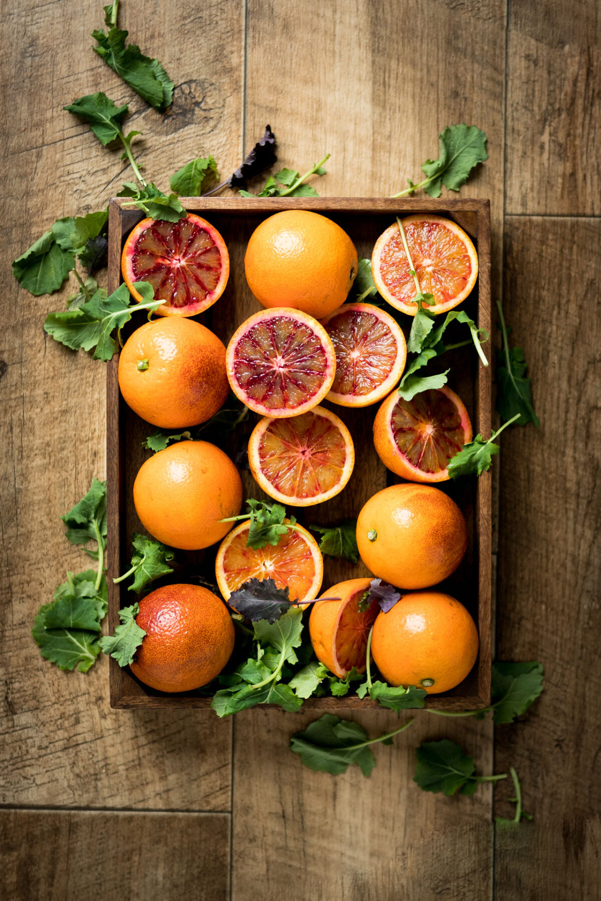 A bunch of blood orange, some whole and some sliced in half, laying in a wooden both on top of a wooden table and garnished with fresh salad greens.
