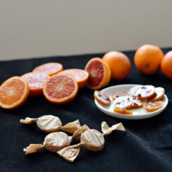 cropped image of toffees on a plate of salted blood orange toffees with fresh orange slices and whole oranges, accompanied by three individually wrapped toffees resembling candies