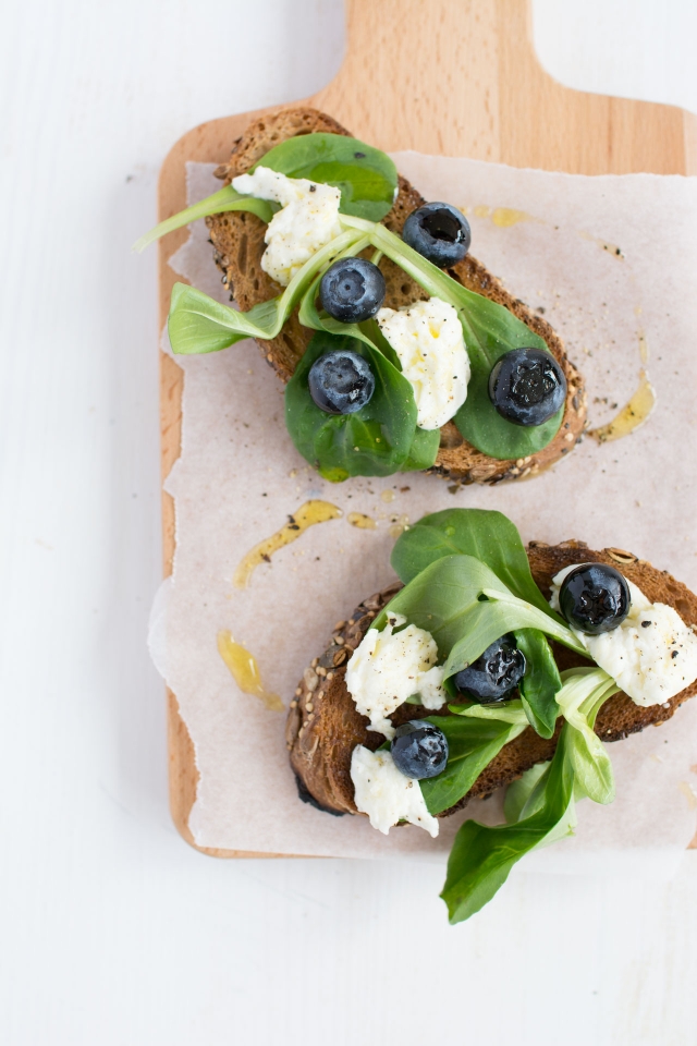 top view image of two slices of bread adorned with fresh blueberries, caprese leaves, and mozzarella, drizzled with olive oil