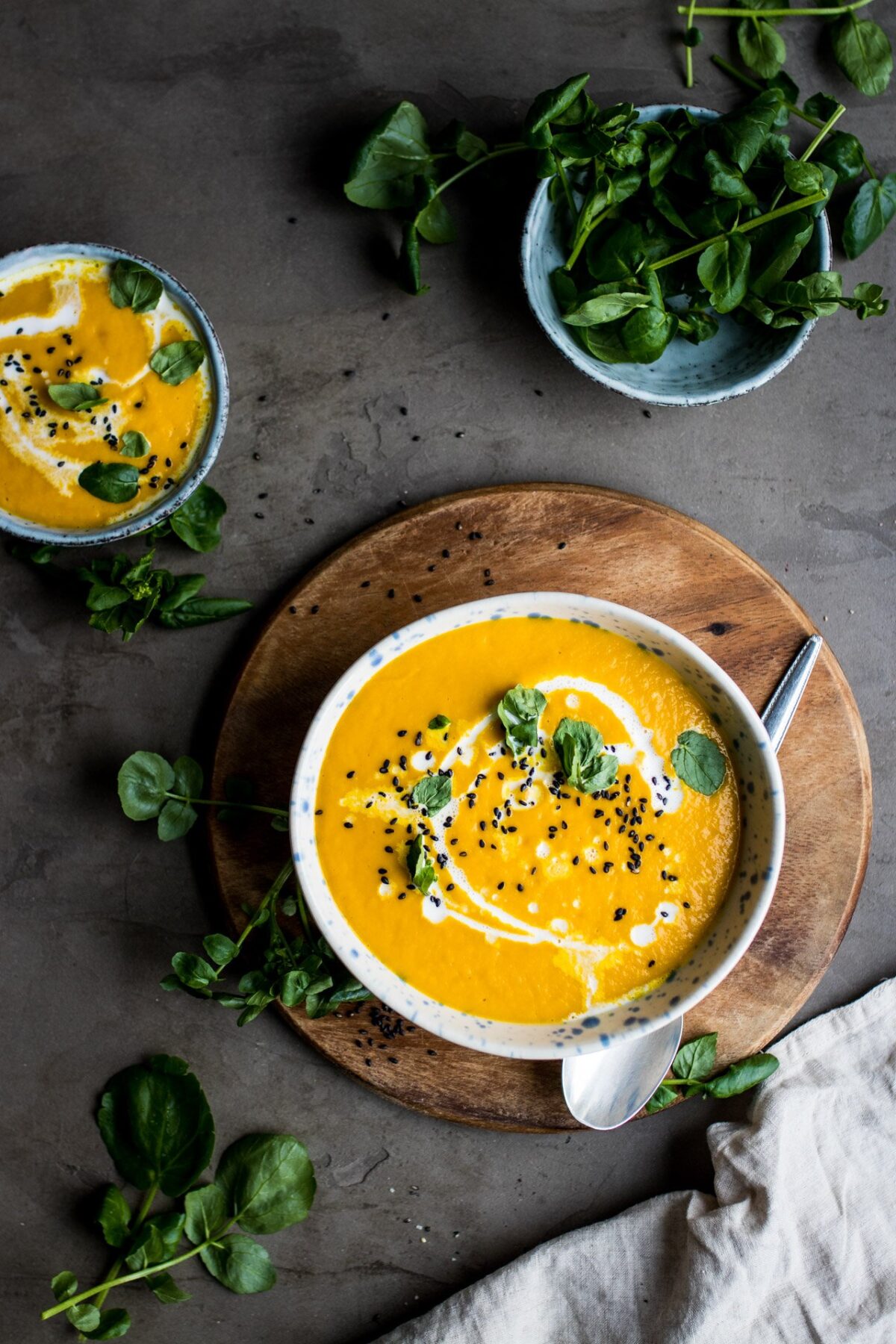 A bowl of turmeric soup with coconut milk drizzled in it. The bowl is on a wooden platter with a smaller bowl of turmeric soup and some herbs.