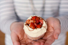 cropped image of a woman holding a single cupcake with fresh cream, strawberries, and balsamic vinegar glaze in both hands