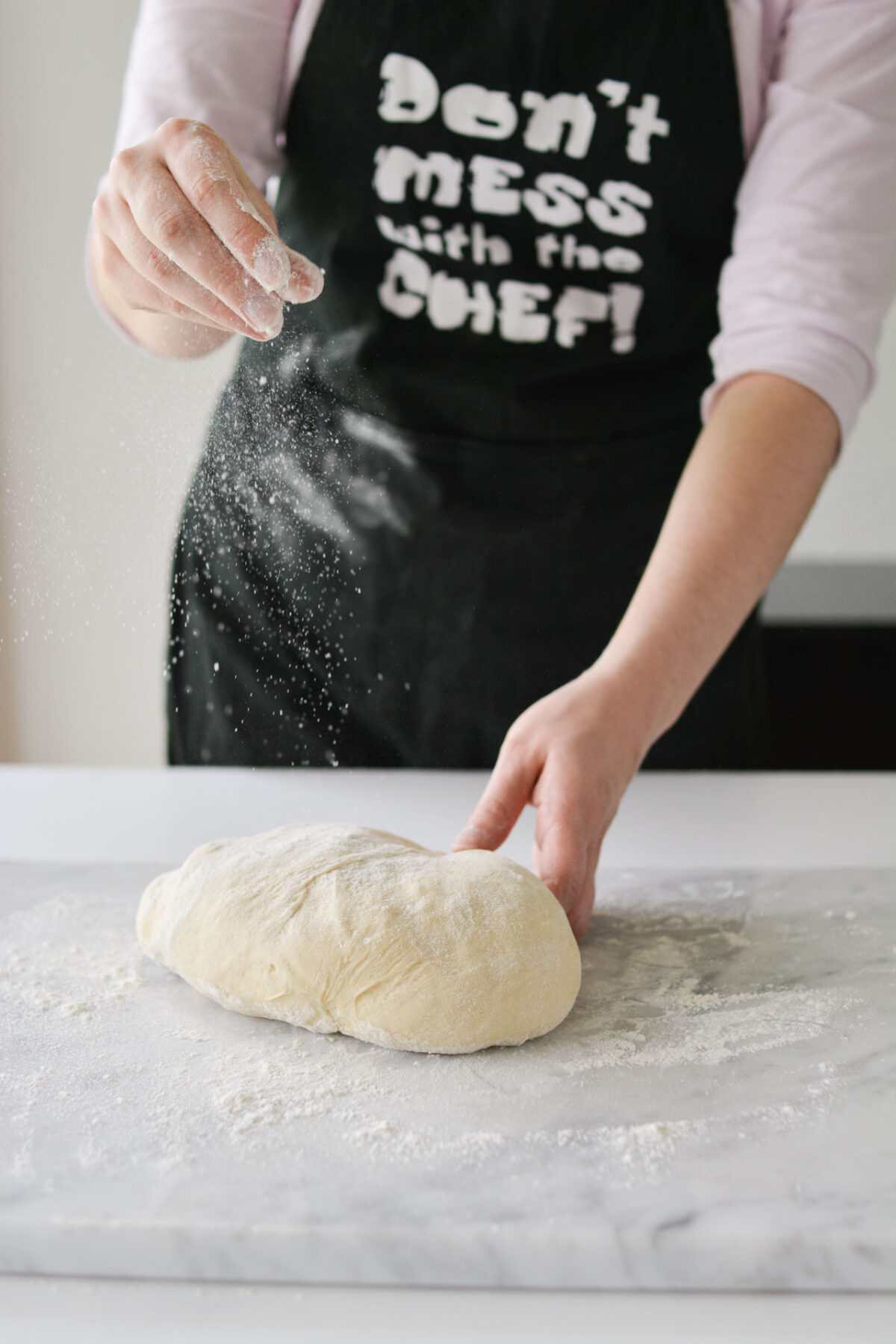 A woman spreading flour onto a kneaded dough on a well-floured surface. Her hands are engaged in the process, creating a rustic and tactile scene of dough preparation.