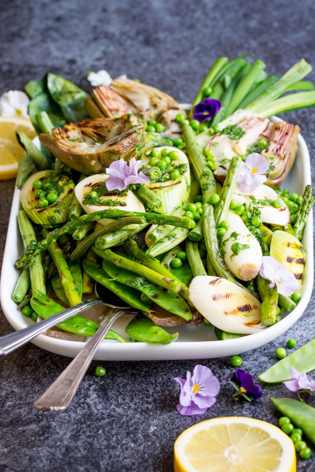 front view image of a serving dish containing grilled spring vegetables including french green beans, spring onions, sugar snap peas, asparagus, leeks, and artichokes, glisten with a homemade lemon parsley butter with fork and spoon
