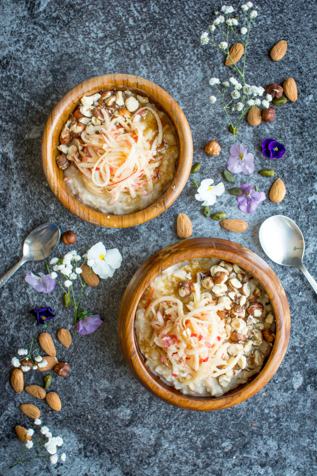 Top-down view of two wooden bowls containing creamy oatmeal, adorned with grated apple, toasted hazelnuts, and a sprinkle of sweet brown sugar.