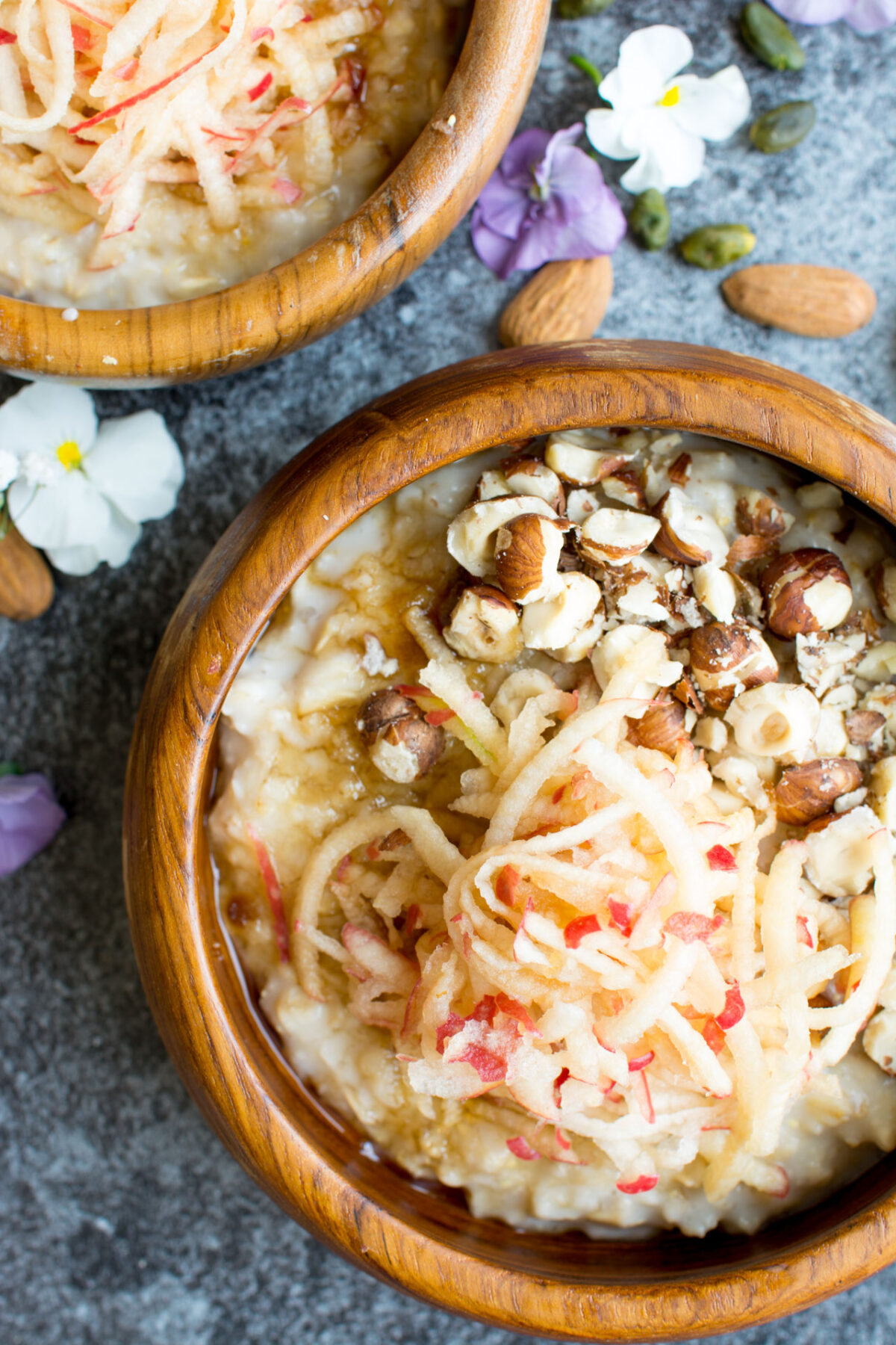 a close-up view image showcases a wooden bowl filled with creamy oatmeal, enhanced with grated apple, toasted hazelnuts, and a delicate dusting of sweet brown sugar.