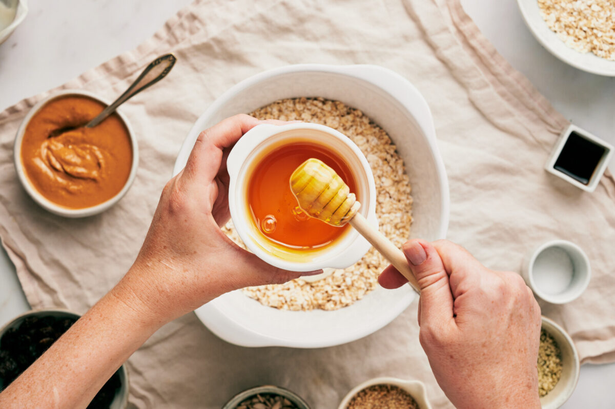 A woman's hand holding a bowl of honey with a honey dipper and other ingredients blurred in the background.