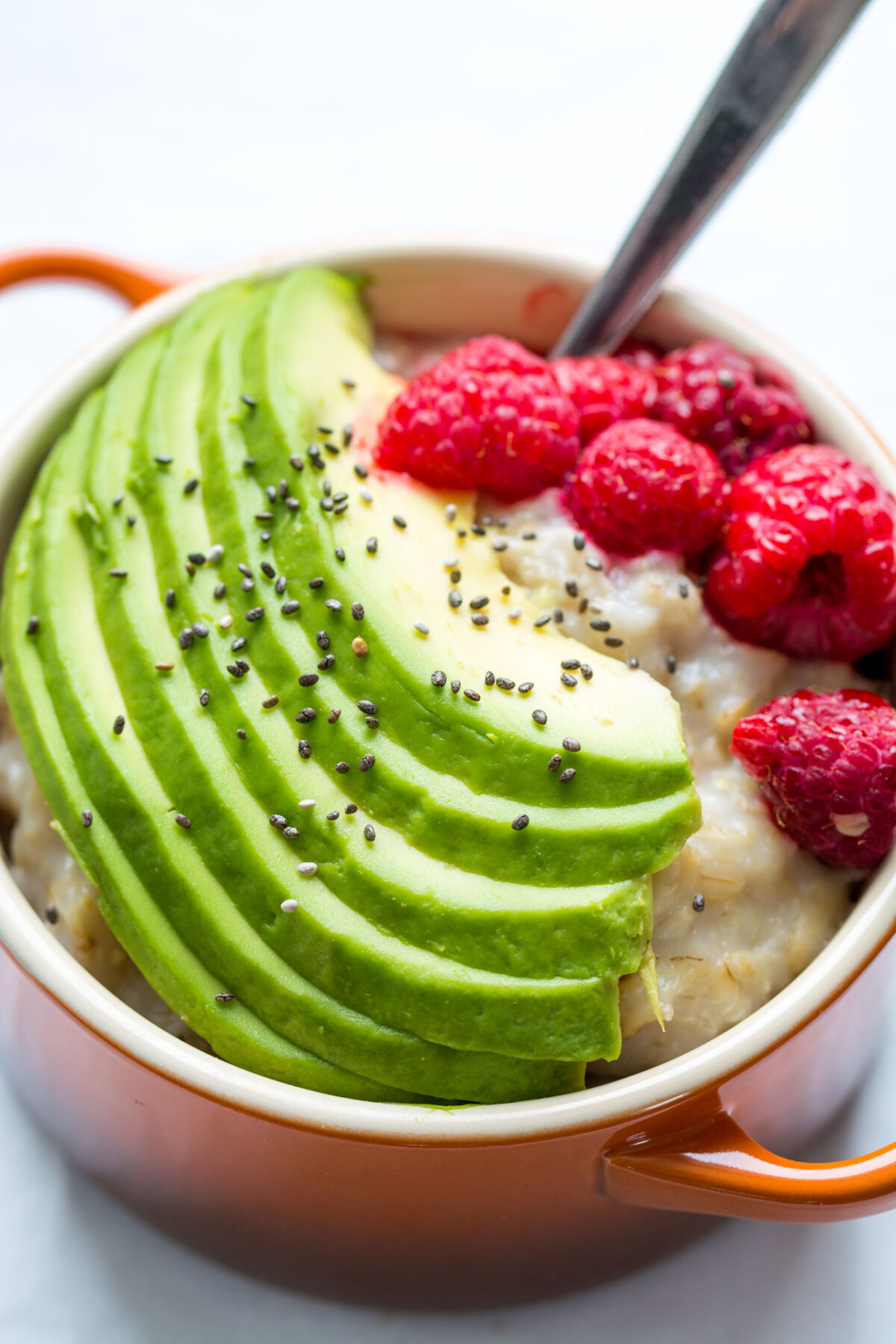 close up image of a bowl containing an oatmeal with slices of avocado, chia seeds, and raspberries
