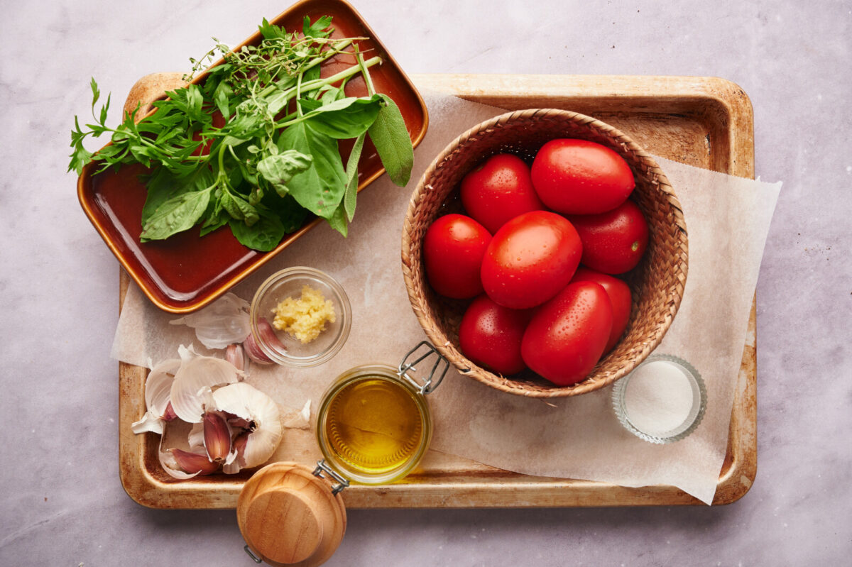 Ingredients for preparing oven roasted tomatoes including plum tomatoes, basil, garlic, olive oil, and salt.