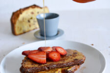 cropped image of a hand drizzling maple syrup onto a plate of panettone french toast, garnished with fresh strawberry slices