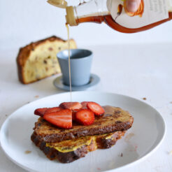 cropped image of a hand drizzling maple syrup onto a plate of panettone french toast, garnished with fresh strawberry slices