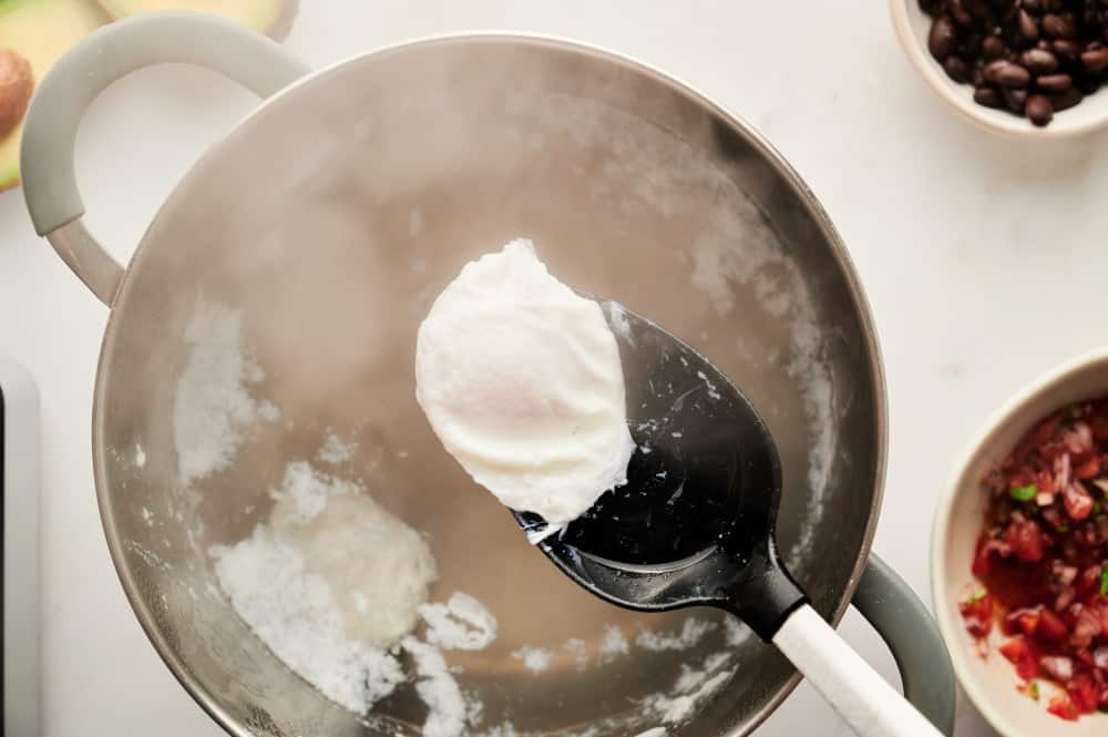 A poached egg being held over a pot of steaming water with a large spoon.