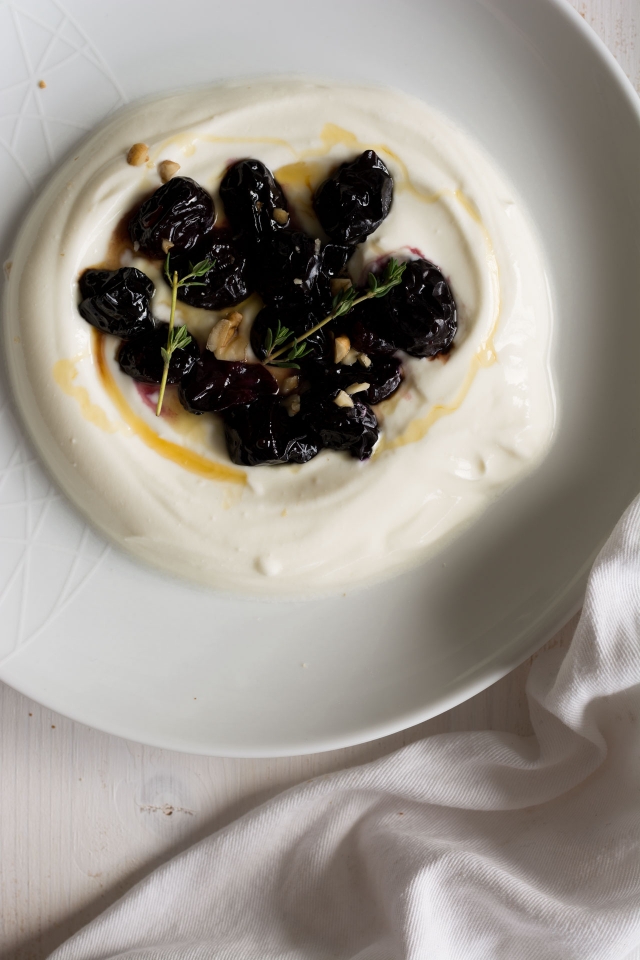 round plate containing with yogurt topped with roasted grapes, and thyme, drizzled with honey and crushed walnuts