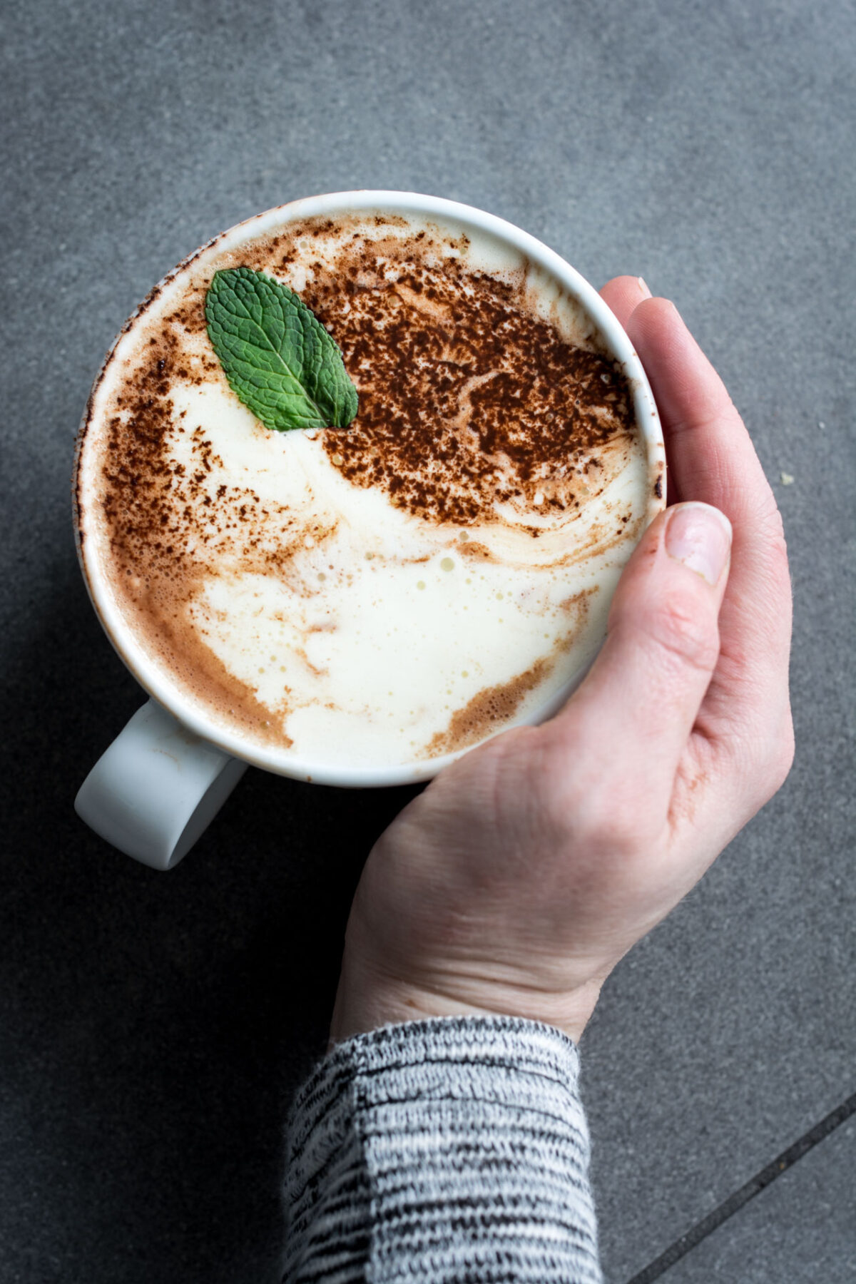 A woman's hand wrapped around a cup of dairy-free peppermint hot chocolate with cocoa sprinkled on top and garnished with a mint leaf.